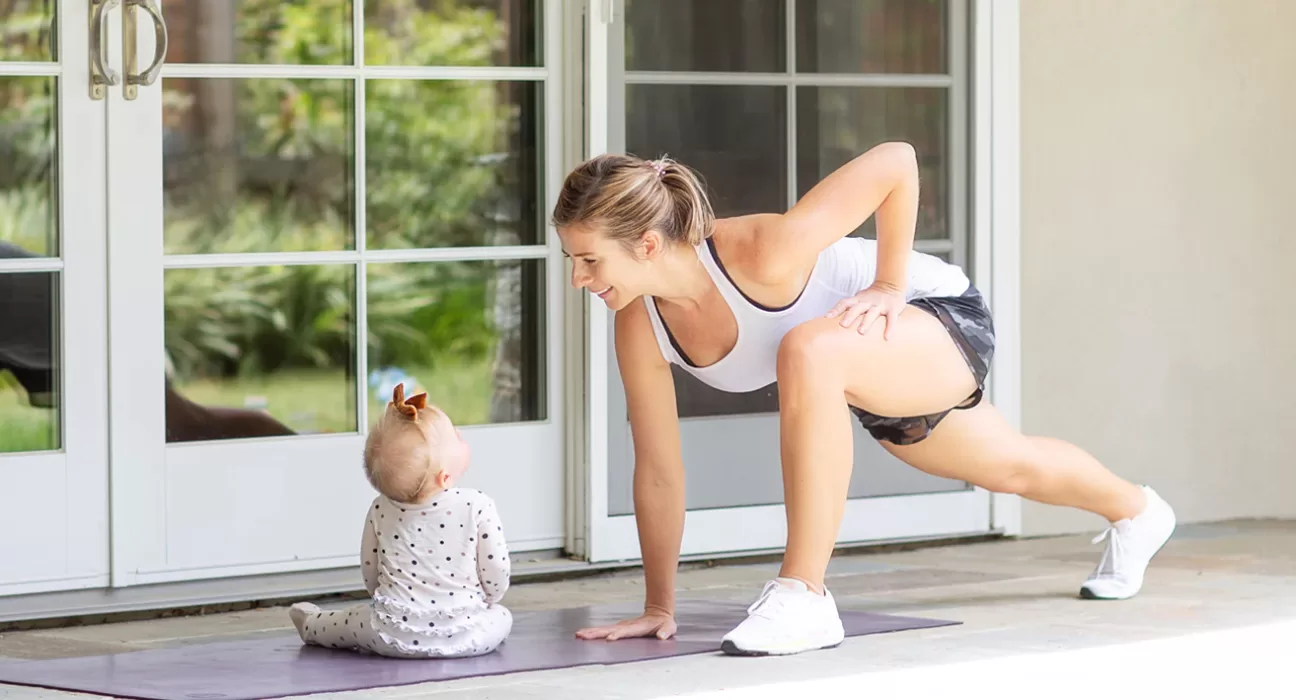 A busy mother doing a no-equipment squat or plank in a living room while a baby or child is playing nearby, emphasizing convenience.