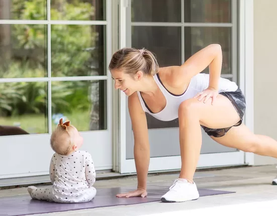 A busy mother doing a no-equipment squat or plank in a living room while a baby or child is playing nearby, emphasizing convenience.
